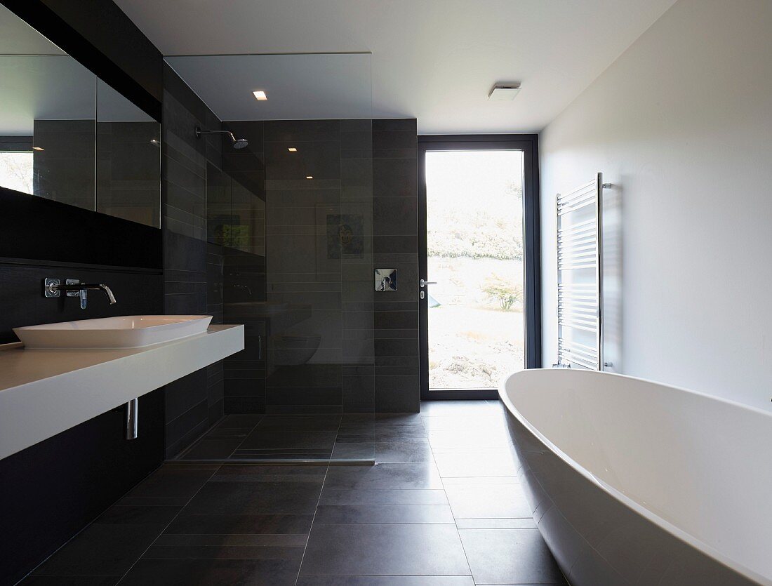 Schwarz-weisses Designerbad mit freistehender Badewanne auf dunklem Fliesenboden, gegenüber weisser Waschtisch und Dusche mit Glastrennwand