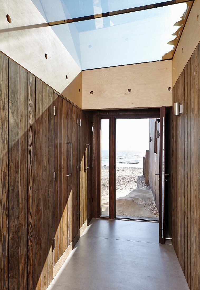 Hausflur mit Einbauschrank aus Holz und grossflächiges Glasdach, Blick durch offene Haustür auf Sandstrand und Meer