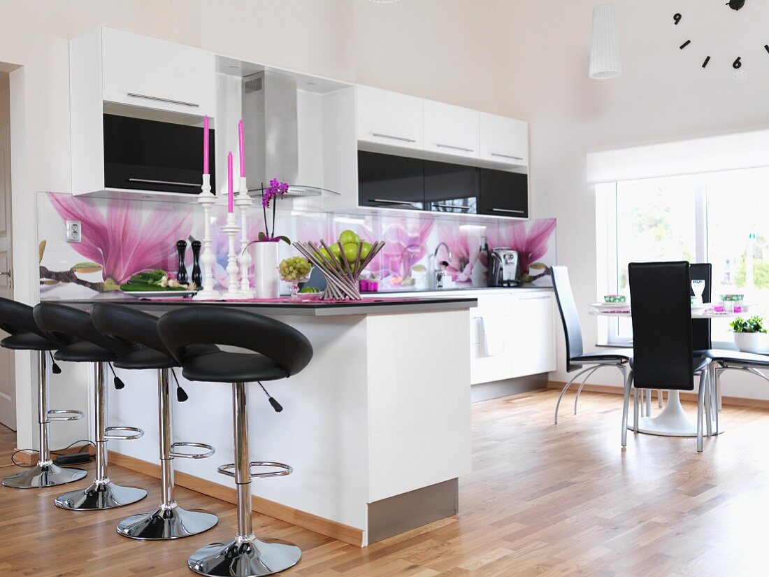 White fitted kitchen with magnolia-patterned splashbacks, leather bar stools at counter and dining area in background next to window in open-plan interior