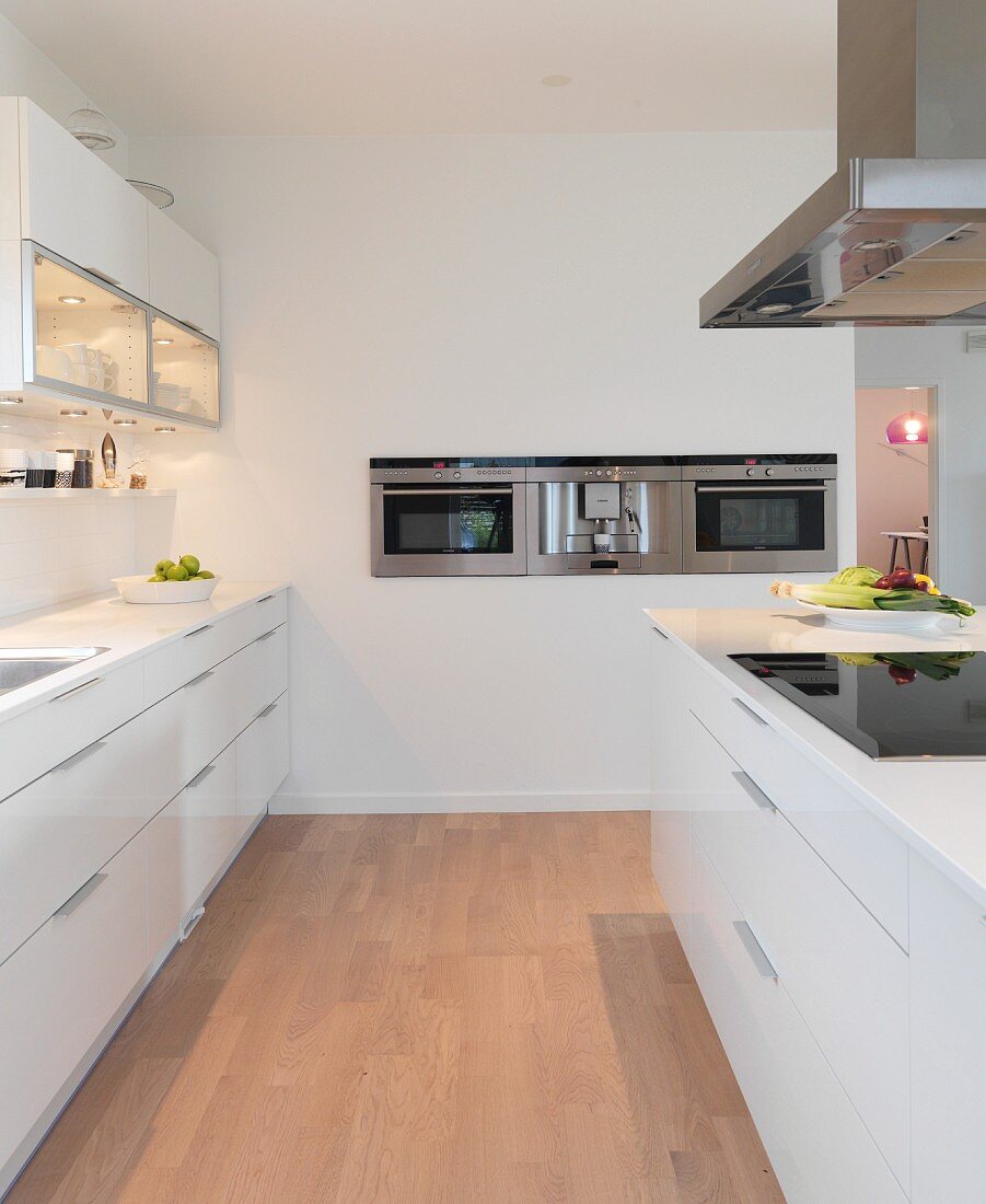 weiße Designerküche mit beleuchtetem Oberschrank, Kochinsel und in die Wand integrierten Edelstahlgeräten