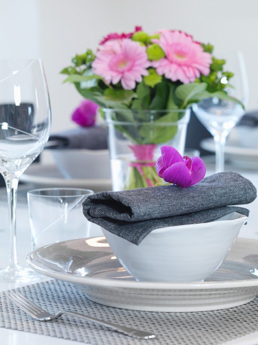Festive place setting with white china crockery and purple flower on grey linen napkin in front of vase of flowers