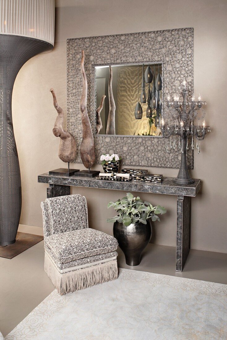Silbergraues Arrangement aus Skulpturen und Leuchter auf Konsolentisch, ornamental gerahmtem Spiegel und Polstersessel mit Fransenbordüre