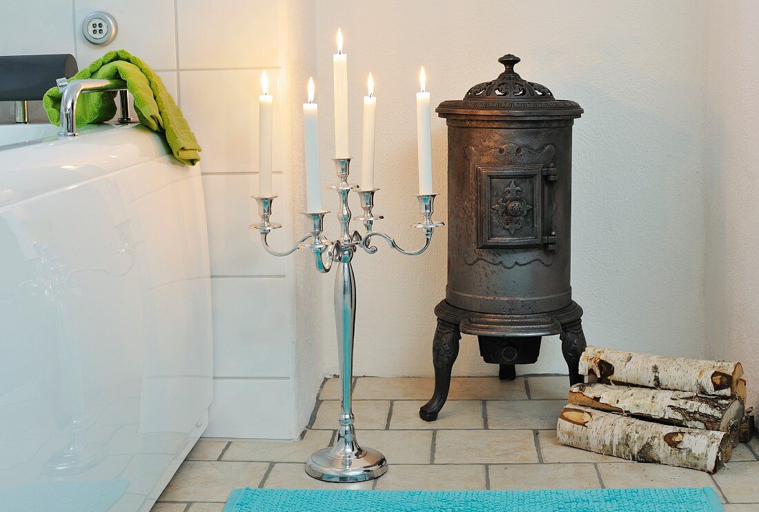 Lit, white candles in floor candlestick, iron stove and small log pile in bathroom