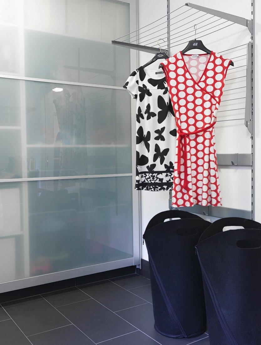 Moderner Waschraum mit schwarzen Wäschebehältern unter Wäscheständer, darauf Damenkleider gehängt, vor Schrank mit Glas Schiebetüren