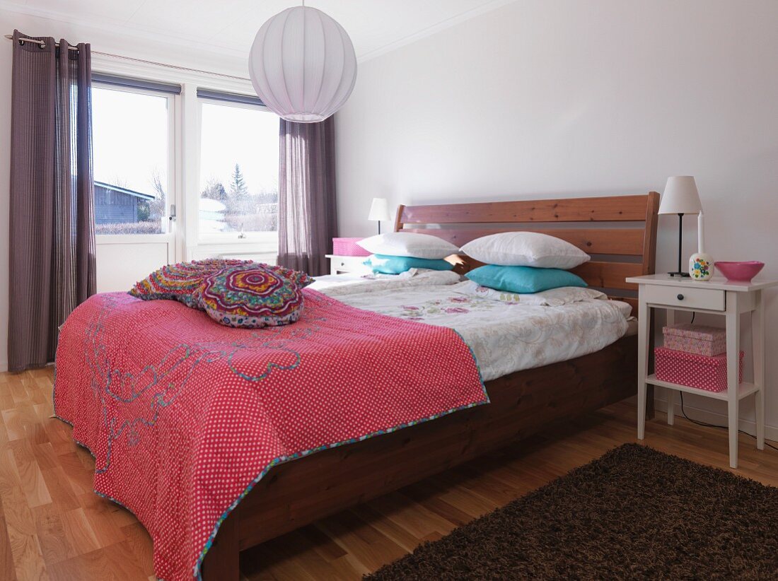 Bett mit Massivholzgestell und Kopfteil, darauf bunte Tagesdecke und Kissen, neben Balkontür im Schlafzimmer