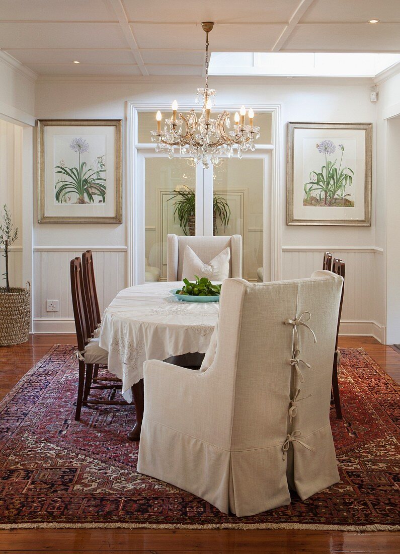 Sessel mit heller Husse und Stühle um Tisch mit weißem Tischtuch, darüber Kronleuchter in traditionellem elegantem Esszimmer