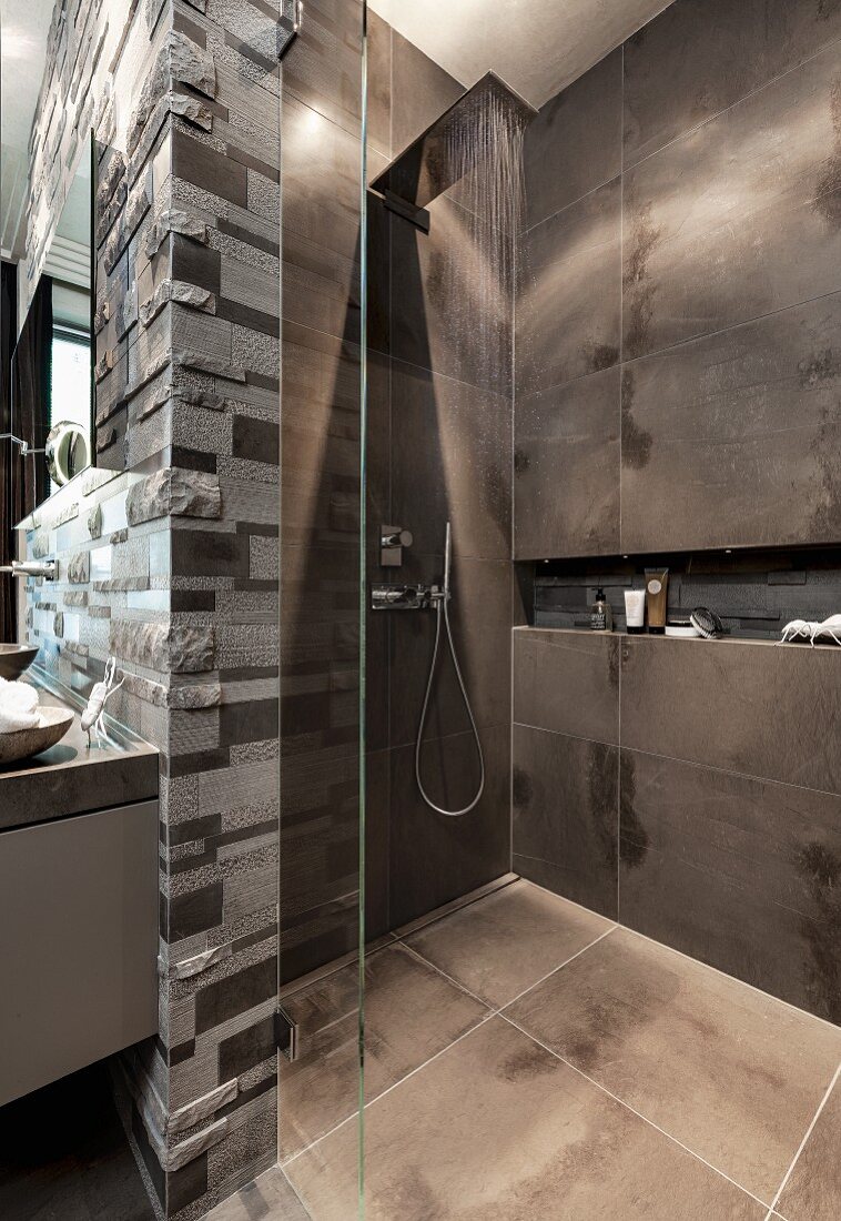 An open rain shower with dark tiles and a glass partition wall with limestone relief tiles behind the sink