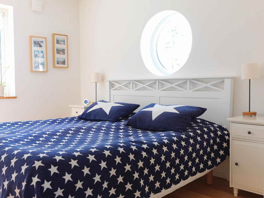 Porthole above double bed in Scandinavian bedroom with blue bedspread and pillows with pattern of stars