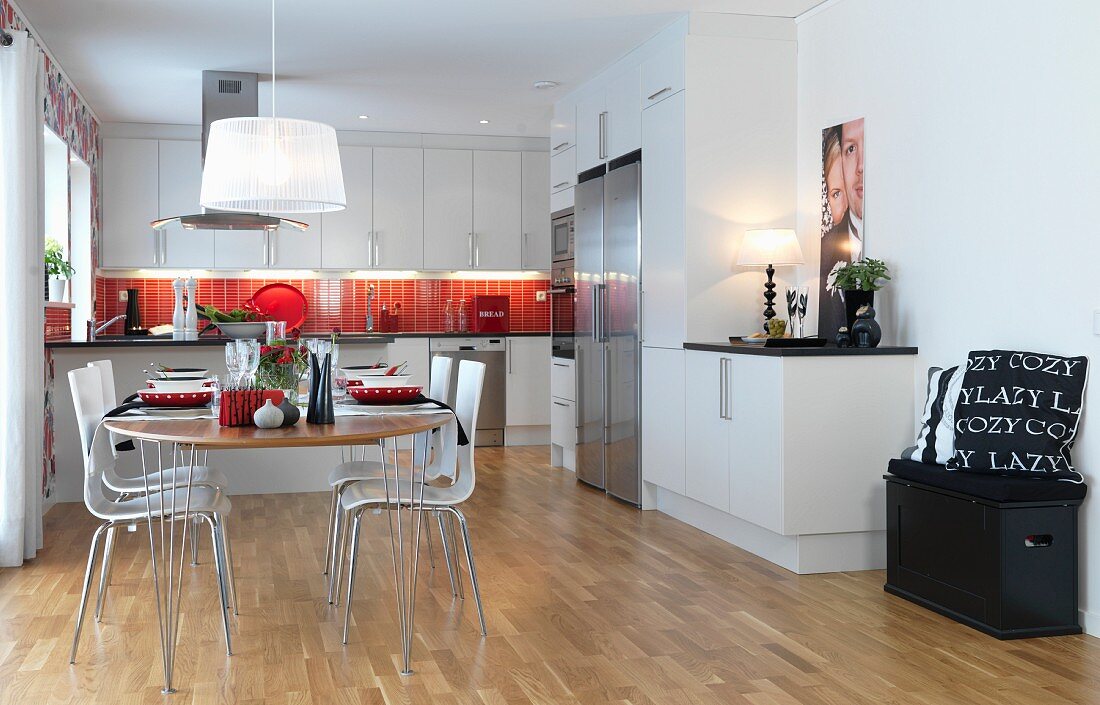 Festively set, oval dining table and shell chairs in open-plan, white, fitted kitchen with red splashback