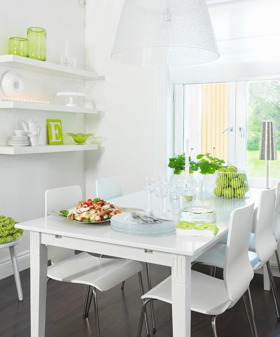 White dining table and chairs below window next to crockery on floating shelves with green accents