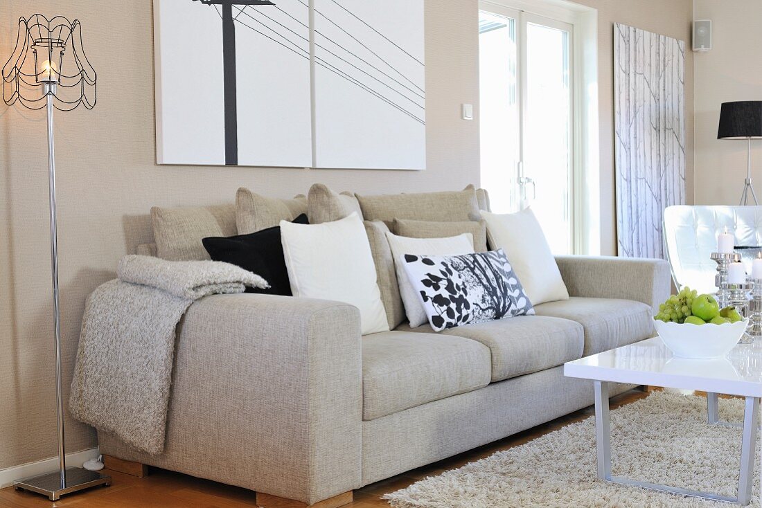 Naturfarbenes Sofa mit Kissen, seitlich Stehleuchte im Retrostil und moderner Couchtisch auf Teppich in zeitgenössischem Wohnzimmer