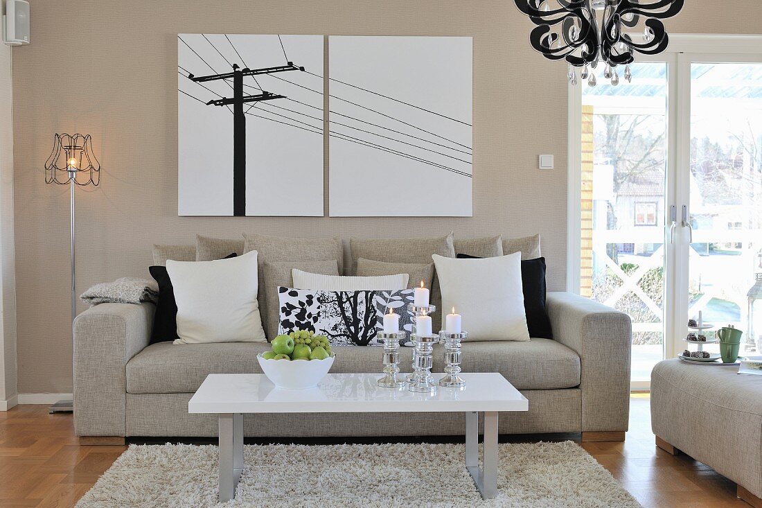 Coffee table on flokati-style rug in front of sofa with row of scatter cushions and retro standard lamp next to modern artwork on wall painted pale grey