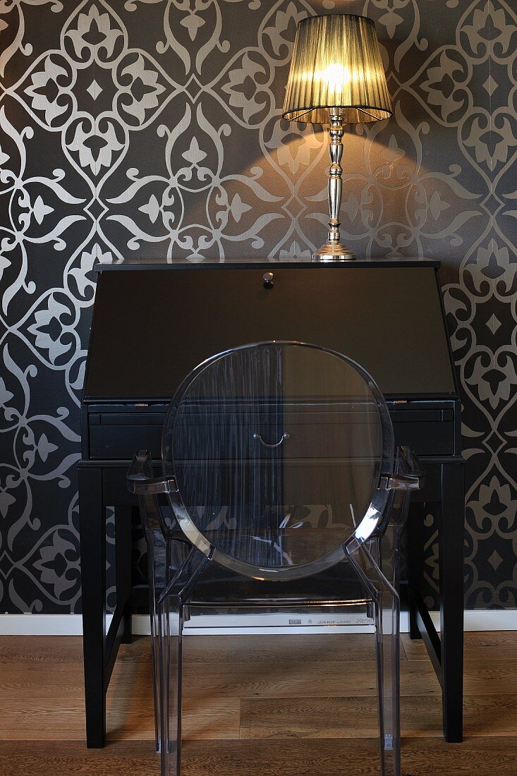 Ghoststuhl vor schwarzem, antikem Sekretär mit Tischleuchte, an Wand schwarze Tapete mit silberfarbenem Ornamentmuster
