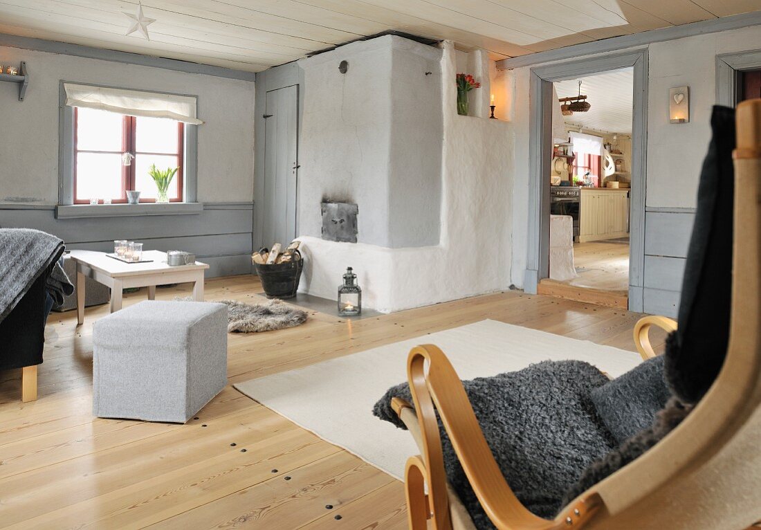 Rustic interior, pouffe and simple coffee table below window; armchair with sheepskin blanket in foreground