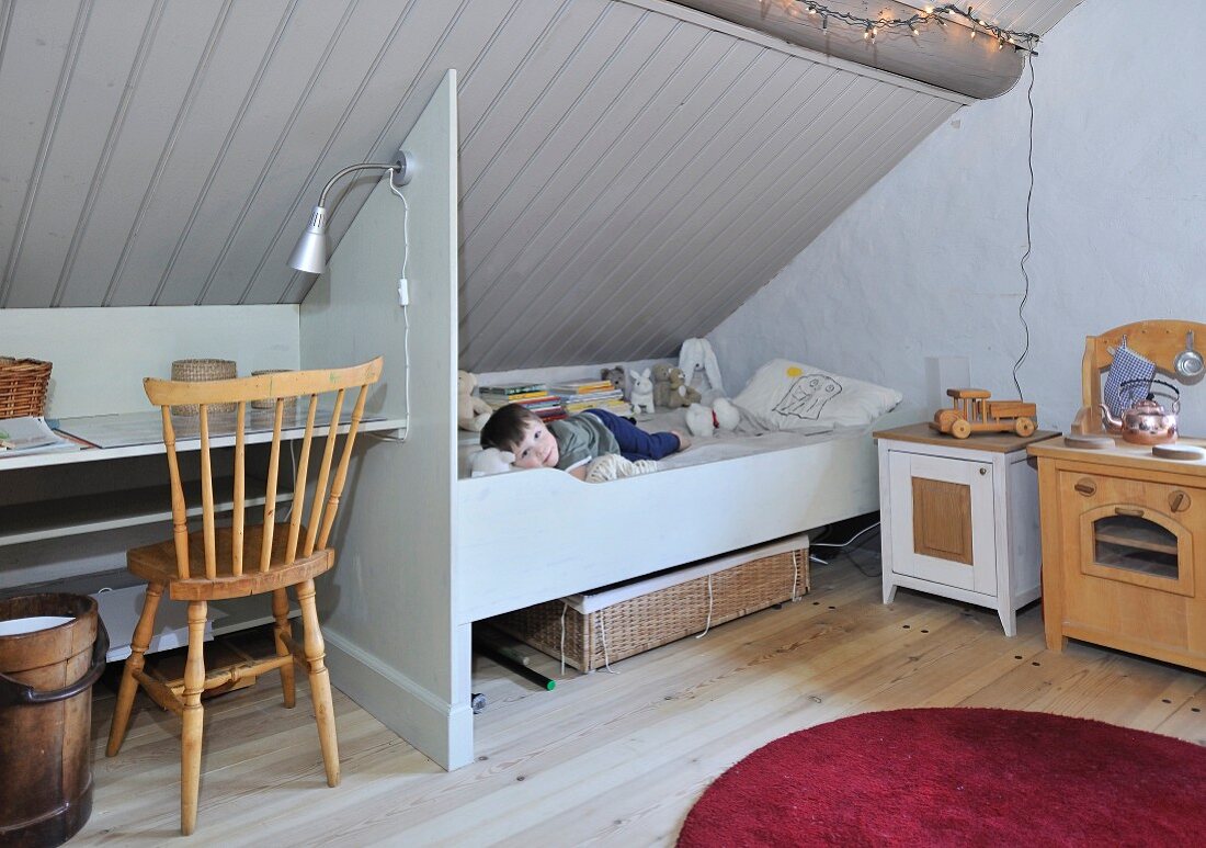 Simple, child's bedroom in attic with little boy in bed below sloping, wood-clad ceiling and partition between bed and desk