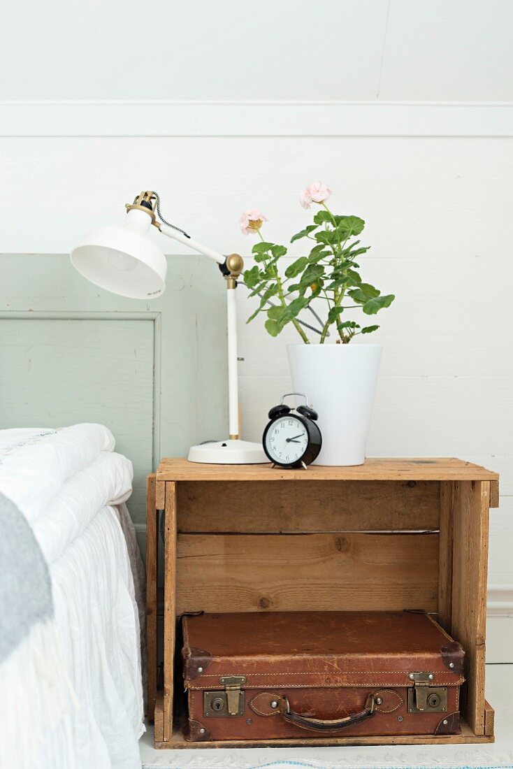 Retro Tischleuchte und weisser Blumentopf mit Geranie auf Holzkiste als Nachttisch, darunter Vintage Lederkoffer, neben Bett