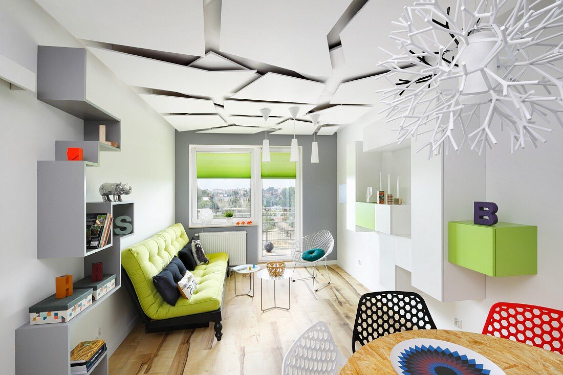 Schmales Wohnzimmer mit hellgrauem Zick-Zack-Regal und grünem Sofa; im Vordergrund Essplatz