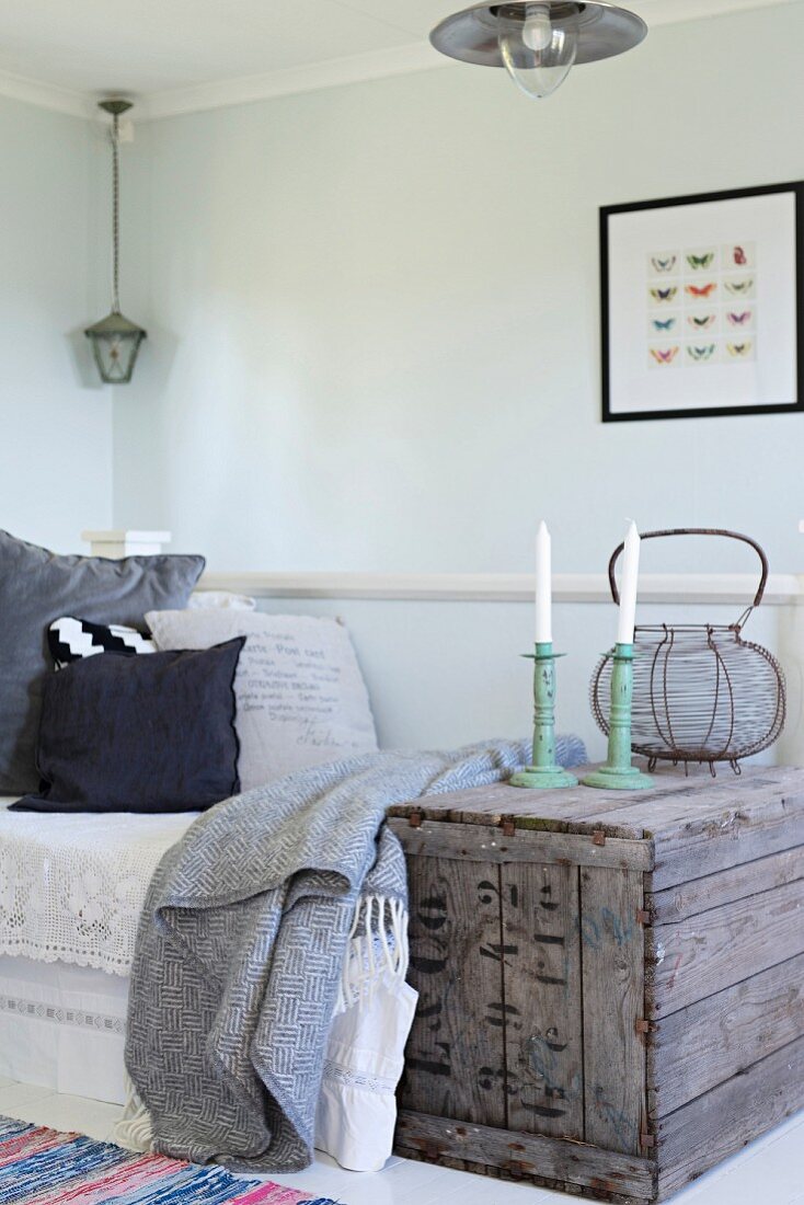 Kerzenhalter mit weissen Kerzen auf Vintage Holzkiste neben Tagesbett in schlichter Zimmerecke
