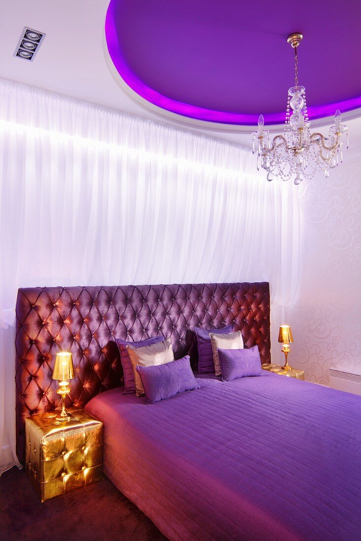 Elegant bedroom in shades of purple, gold bedside lamps on gold pouffes, purple ceiling panel and crystal chandelier