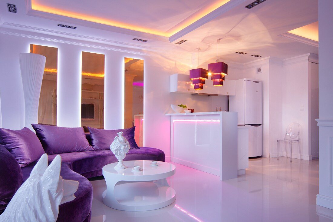 Lila Polstersofa und weißer, runder Designer-Couchtisch in offenem Wohnraum mit stimmungsvoller Beleuchtung