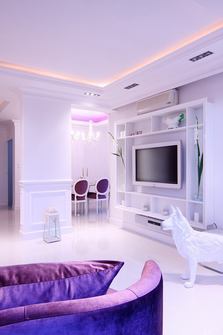 Open-plan, elegant, white living area with purple accents, indirect lighting and plasma TV on open-fronted shelves