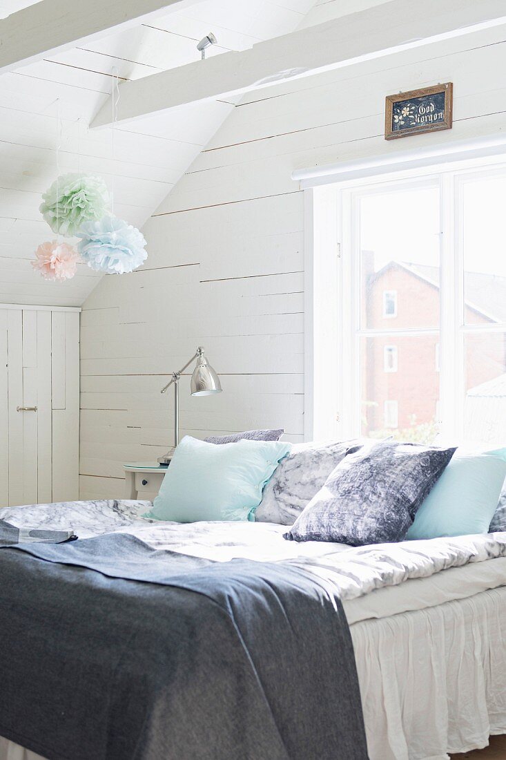 Kissen und Decken auf Bett vor Fenster in holzverkleidetem Dachzimmer; Deko mit pastellfarbenen Papierpüscheln