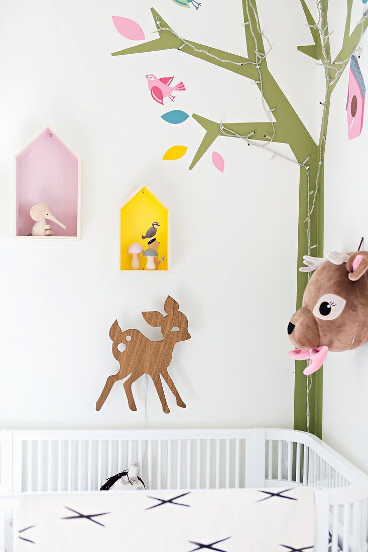 Babybett in Zimmerecke mit Walddeko: stilisierte Baumdarstellung mit Lichterkette, Rehfiguren und Deko-Wandkästen