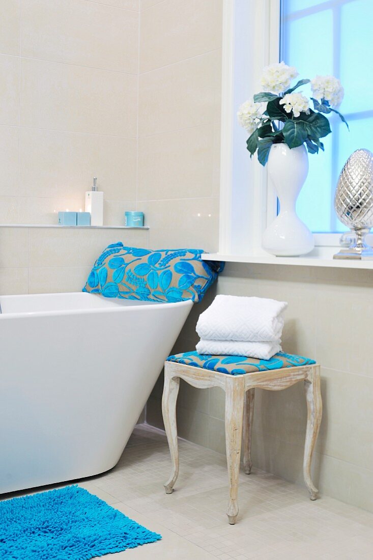 Designer bathtub in bathroom with window, beige tiles, romantic accessories and azure blue, floral devoré textiles