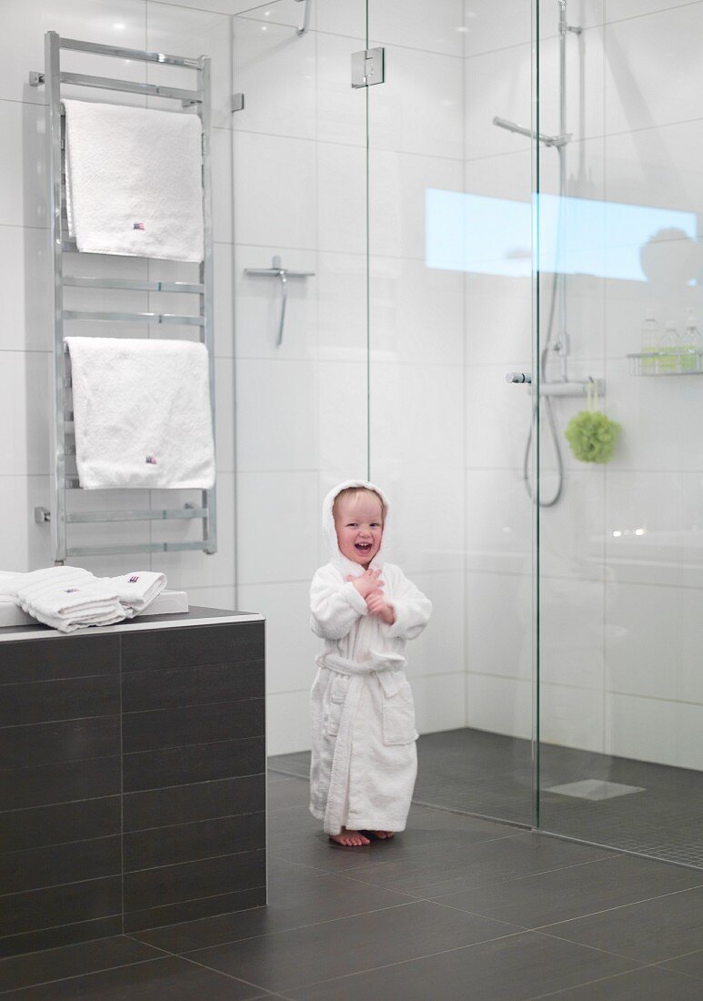 Kleines Kind im Bademantel vor der Glasabtrennung einer ebenengleichen Dusche, Handtuch-Heizkörper im Hintergrund