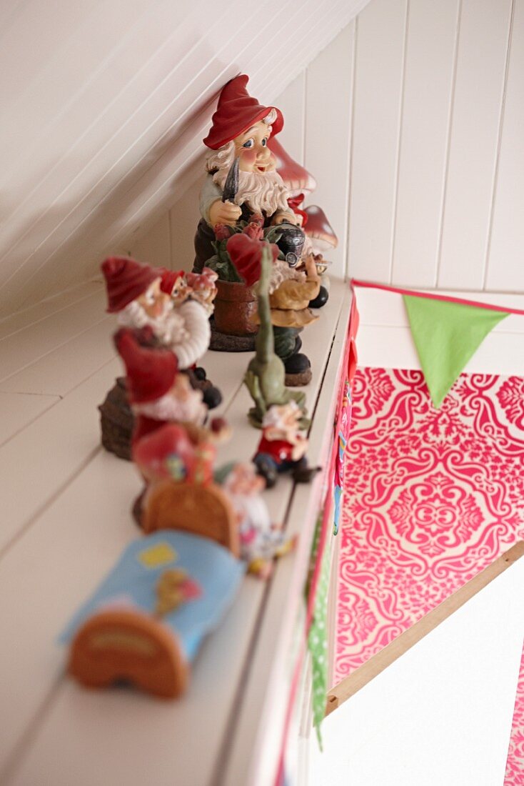 Row of gnomes on shelf on wood-clad sloping wall, colourful bunting and floral pattern in background