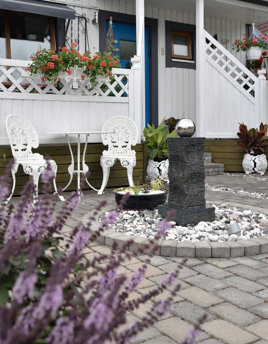 Ziersäule mit Edelstahl Kugel in kreisförmiger Einfassung, im Hintergrund weiße gusseieserne Gartenstühle mit Tisch, auf Terrasse eines weissen Holzhauses