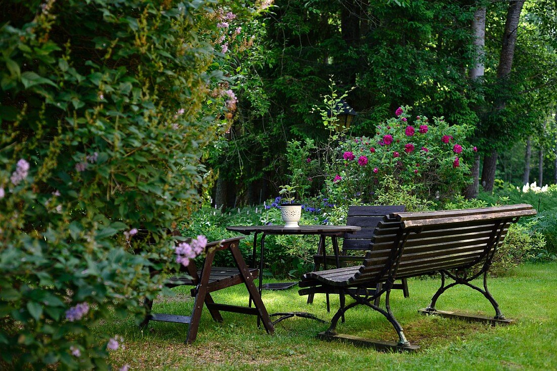 Park bench, table and wooden chairs on lawn in park-style garden with flowering perennials