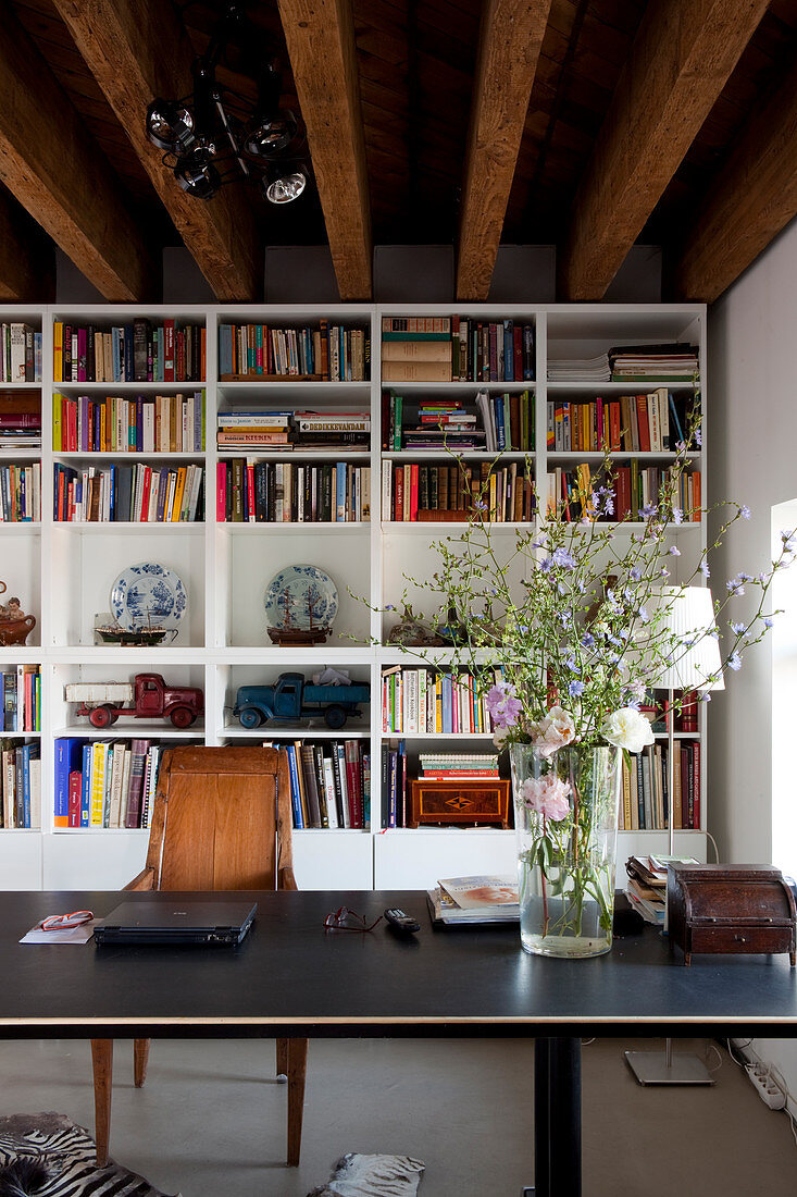 Vase of wildflowers on desk in front of white bookcase in loft apartment with rustic wood-beamed ceiling
