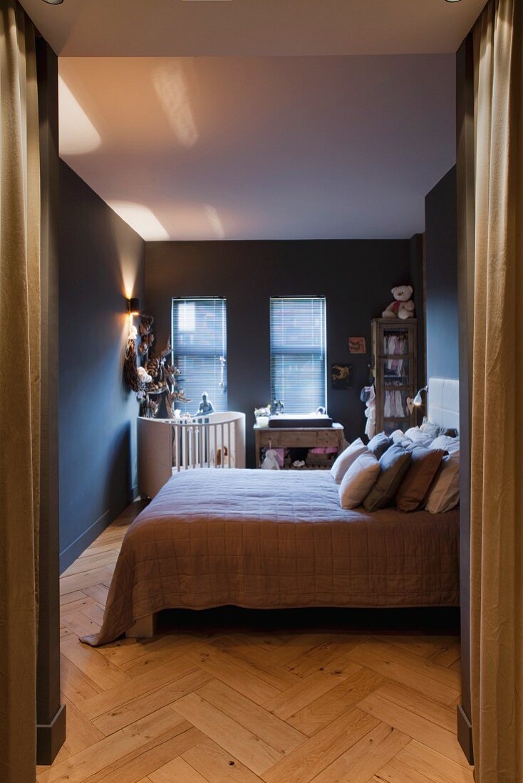 Blick auf Doppelbett mit Kissen, im Hintergrund Kinderbett in dunkelgrau getöntem Schlafzimmer