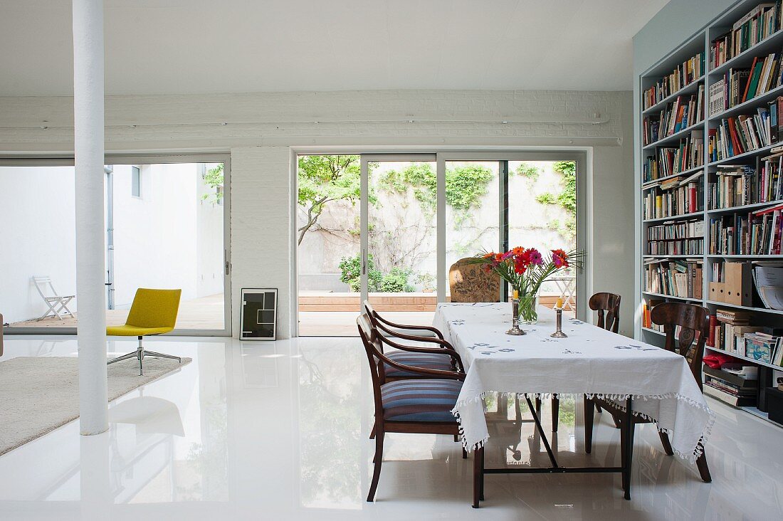 Table with white tablecloth and armchairs opposite bookcase in loft apartment: terrace doors with view into courtyard in background