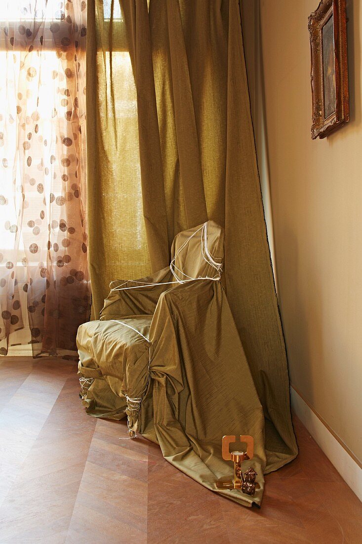 Armchair in corner wrapped and tied in fabric in front of floor-length curtain of matching material