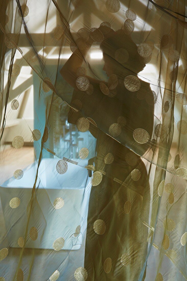 Verschleierter Blick durch transparenten Vorhang auf Frau und freistehende Badewanne im Dachgeschoss