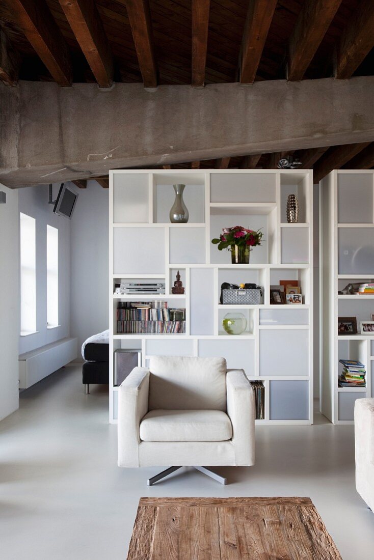 Modern, pale beige armchair in front of white designer shelves with open-fronted and closed modules used as partition in loft apartment