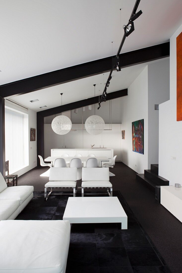 White designer furniture and low coffee table on black rug with dining area and kitchen in background in loft apartment