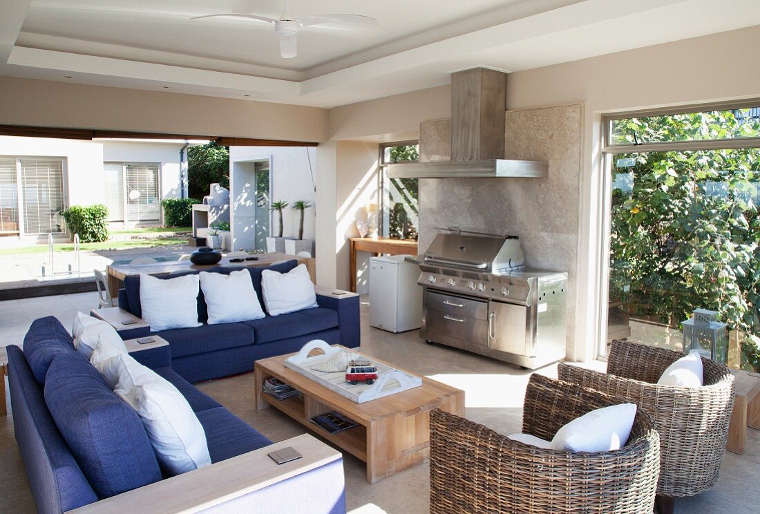 Blue sofas and rattan armchairs in sunny seating area with barbecue; view of pool in courtyard in background