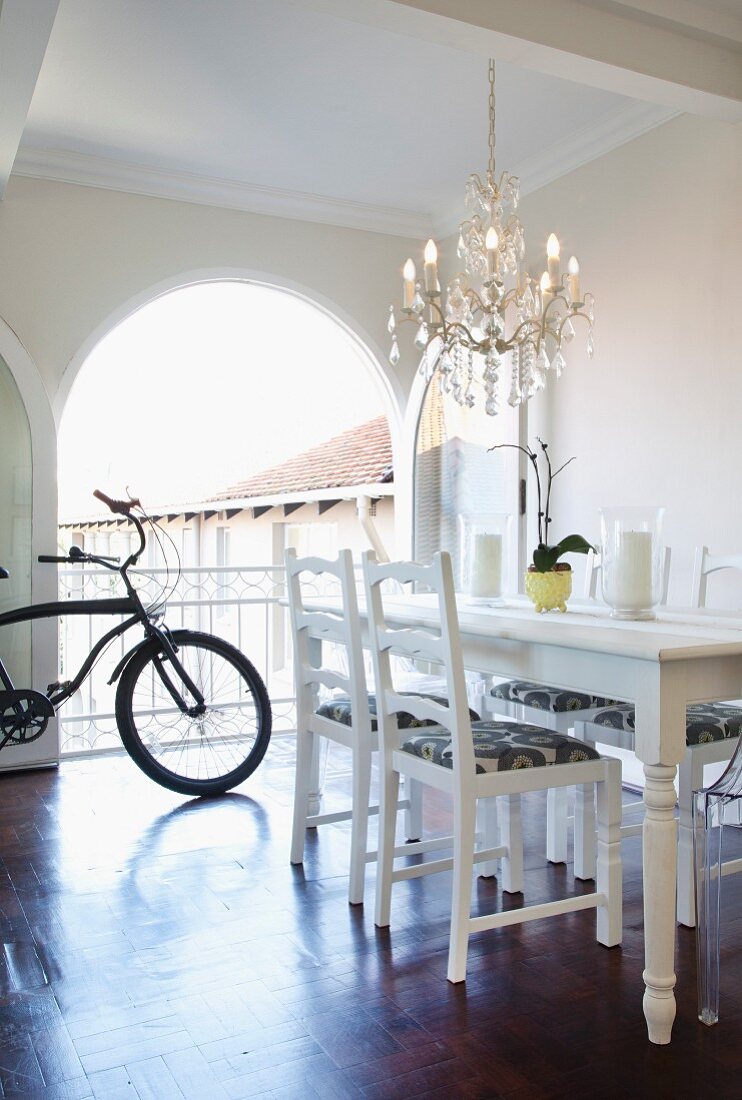 White dining table and refurbished second-hand chairs below chandelier; bicycle in background in front of open arched terrace doors