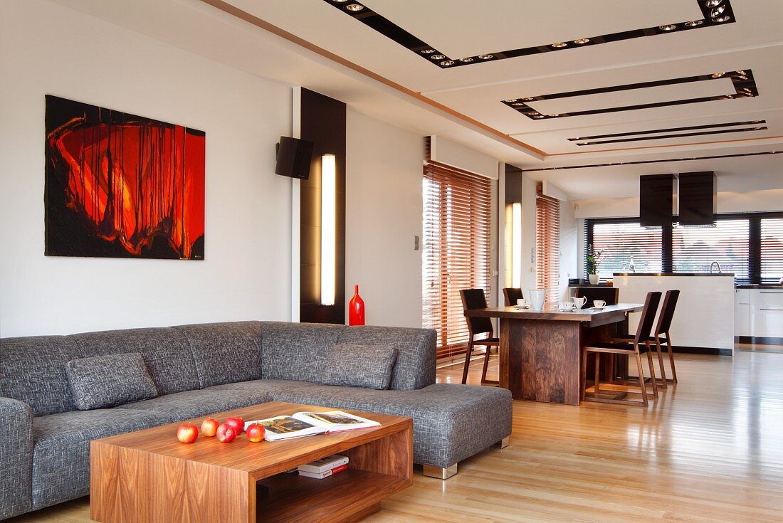 Loungebereich mit grauem Sofa übereck und Coffee Table aus Nussholz, im Hintergrund Essplatz in loftartigem Wohnraum