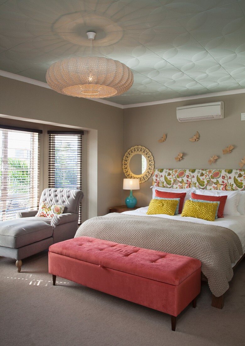 Samtrosa Kleiderbank vor Doppelbett mit Dekokissen und floral gemustertem Kopfteil, Ruhesessel vor Fenster