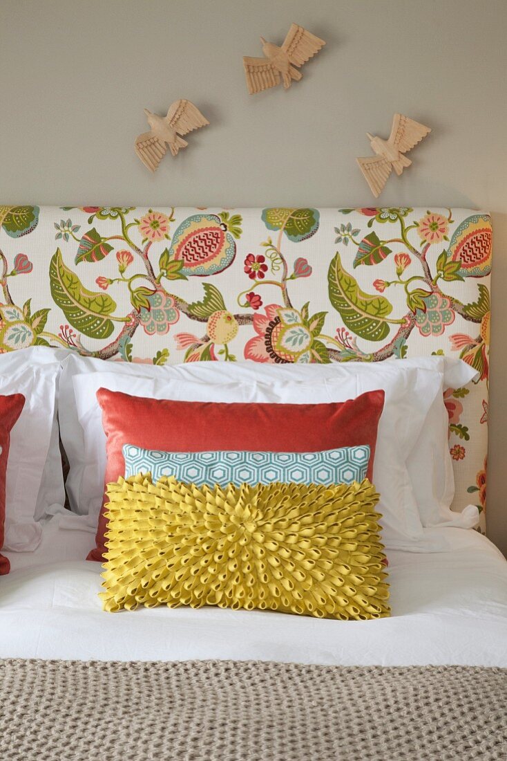 Scatter cushions on double bed with floral headboard below wooden bird figurines on wall