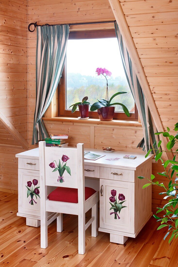 Small desk and chair painted with floral motifs below window in wood-clad attic room