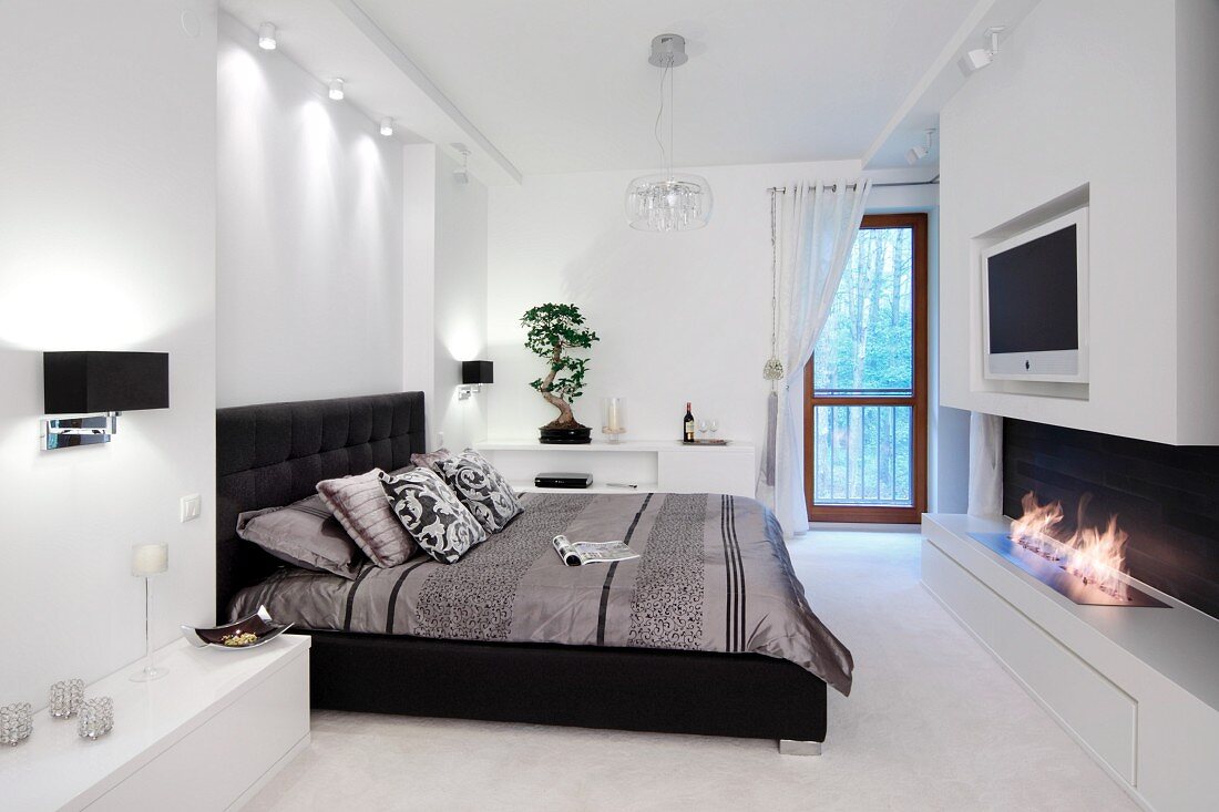 Elegantes Schlafzimmer in Schwarz-Weiß, Doppelbett mit schwarzem Lederbezug gegenüber gasbefeuertem Kamin in weißem, Lowboard