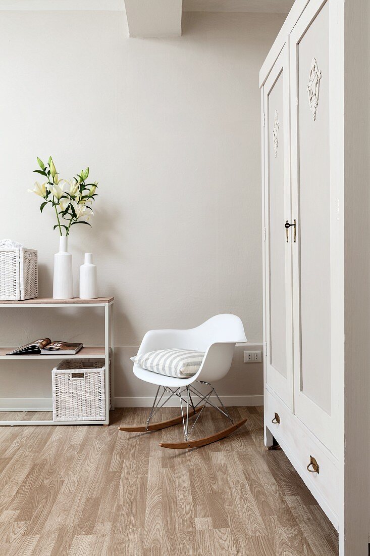 White, designer rocking chair next to painted wardrobe and vase of lilies on top of open-fronted shelving in corner of bedroom