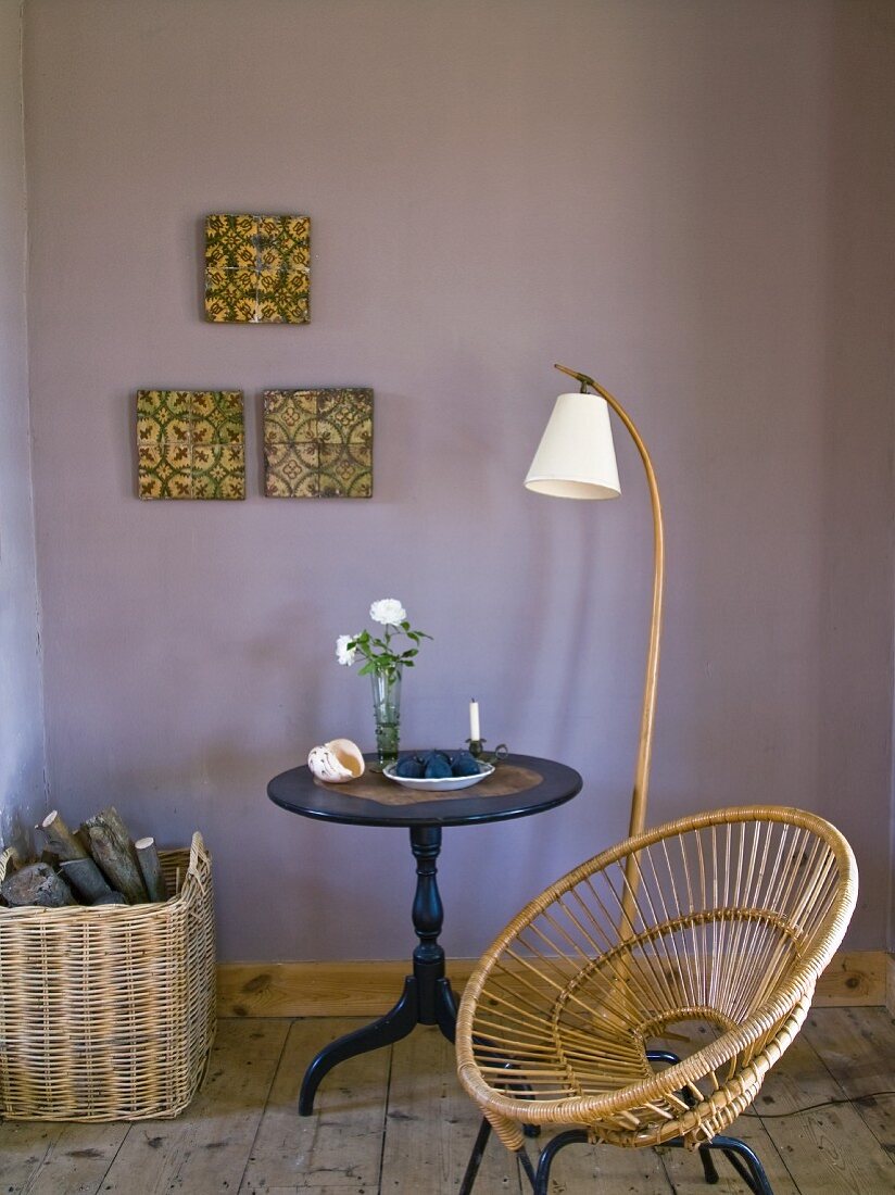 Stuhl aus Geflecht neben Stehleuchte und Bistrotisch vor mauvefarbener Wand mit aufgehängten Vintage Fliesen