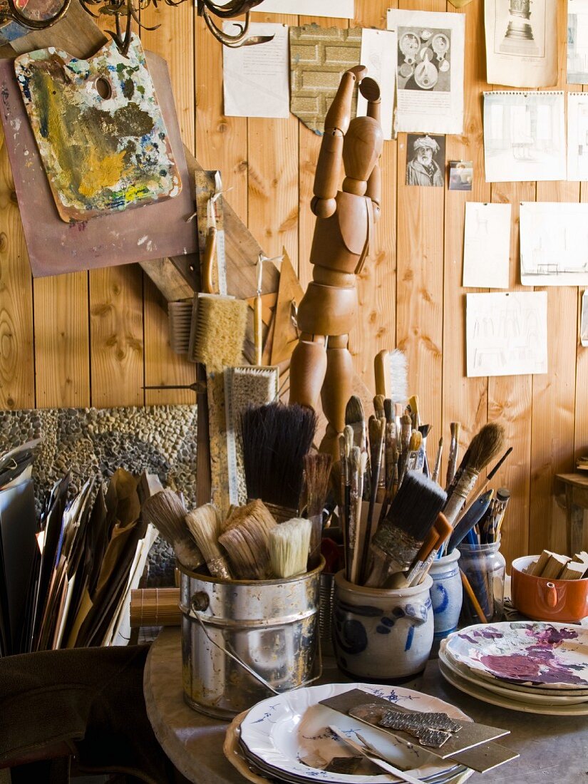 Malutensilien, Behälter mit verschiedenen Pinseln und Holz Gliederpuppe vor Holzwand in einem Atelier