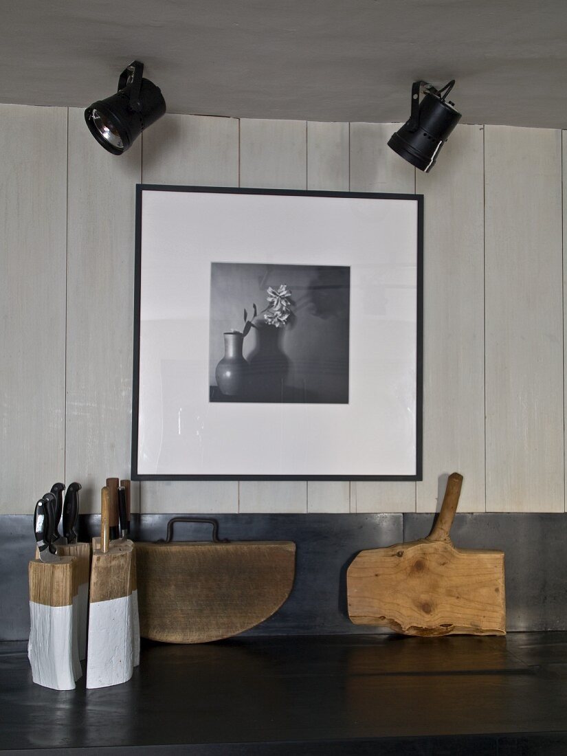 Messerblock und rustikale Schneidebretter auf Küchenarbeitsfläche, Deckenstrahler vor Schwarzweissfoto an weisser Holzwand